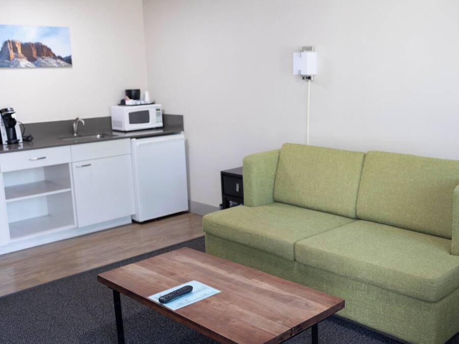 Una sala de estar moderna con un sofá verde, una mesa de centro de madera, una kitchenette blanca con microondas y un cuadro en la pared.