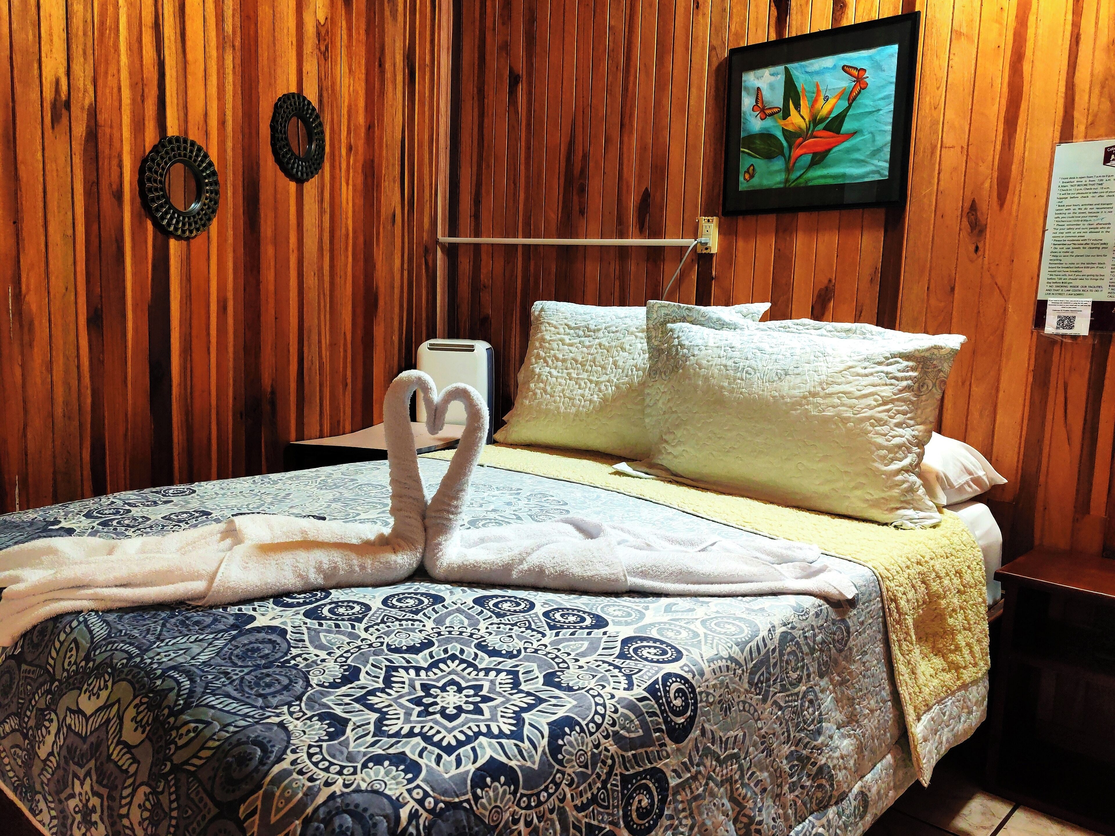 Habitación de hotel con pared de madera, cama con colcha estampada, dos toallas con forma de corazón sobre la cama, un cuadro en la pared y una mesita con lámpara.