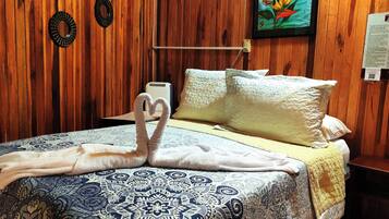 Habitación de hotel con pared de madera, cama con colcha estampada, dos toallas con forma de corazón sobre la cama, un cuadro en la pared y una mesita con lámpara.