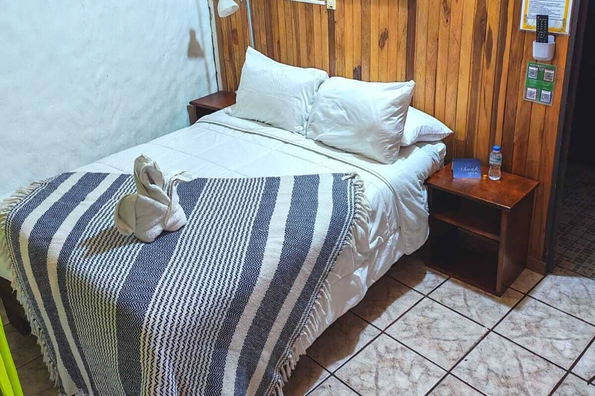 Habitación de hotel con pared de madera, cama con ropa blanca, manta a rayas, mesita de noche con una botella de agua y un cuadro.