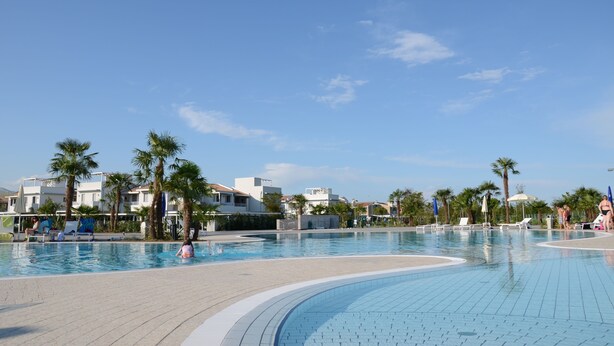 A swimming pool area with lounge chairs, palm trees, and modern buildings in the background. - Villaggio Laguna Blu (Caorle)