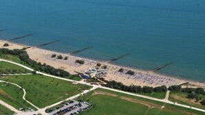 An aerial view of a beach with rows of umbrellas and sun loungers, a parking area, and a small building near the water. - Villaggio Laguna Blu (Caorle)