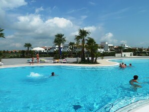 A swimming pool with people swimming and lounging, surrounded by palm trees and buildings. - Villaggio Laguna Blu (Caorle)