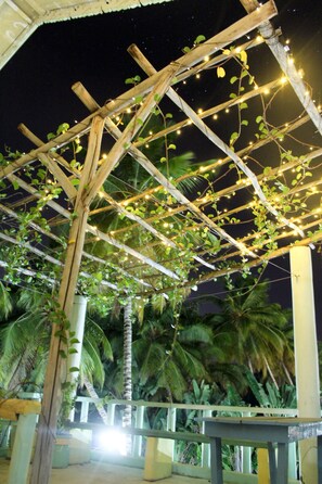 Una estructura de pérgola de madera con luces colgantes y plantas trepadoras, rodeada de palmeras.