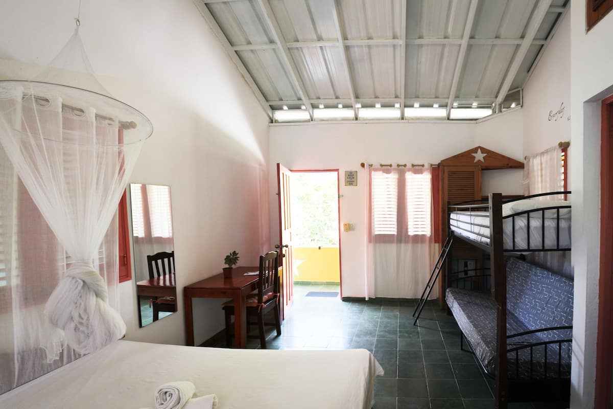 Une chambre comprenant un lit, un bureau, une chaise, une moustiquaire, un lit superposé et une porte donnant sur un espace extérieur.