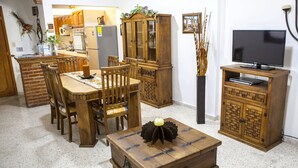 Un comedor con muebles de madera, un televisor sobre un soporte y una cocina al fondo.