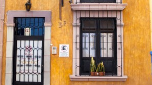 Un edificio amarillo con un toldo verde, una puerta negra con un letrero y dos ventanas con macetas.