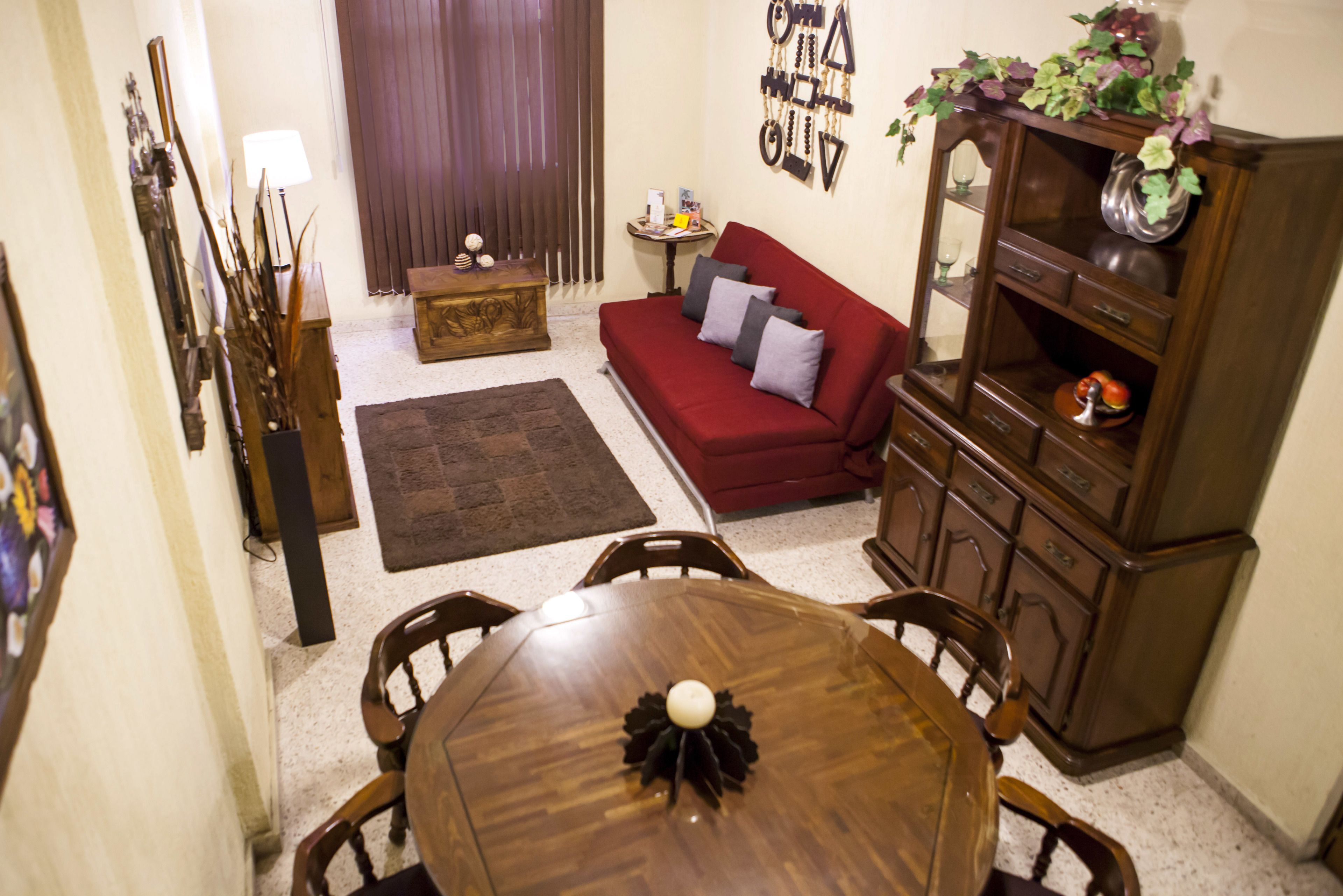 A dining area with a wooden table and chairs, a red sofa, a small side table, and a cabinet with decorative items.