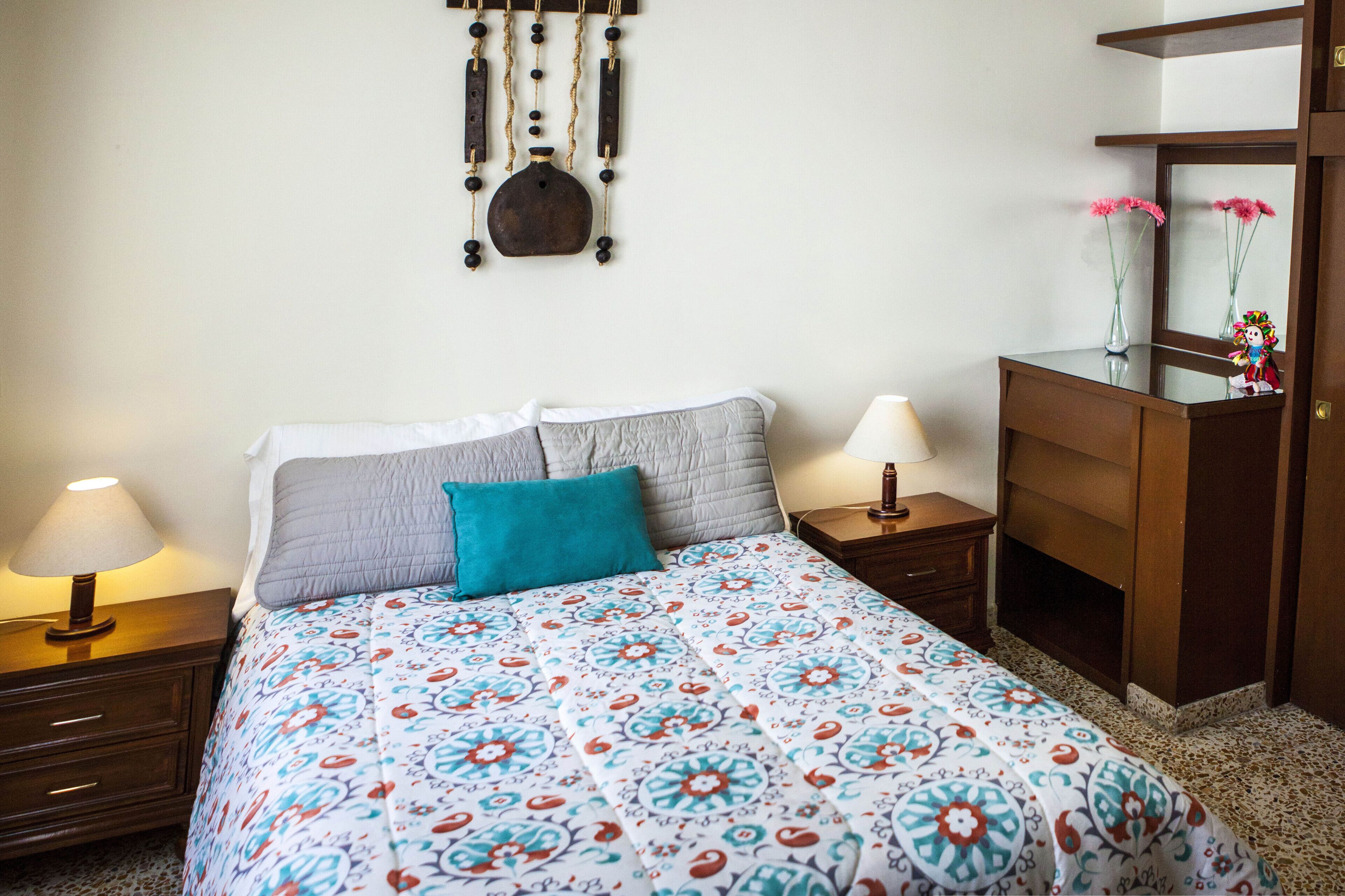 A bedroom with a bed, two bedside tables with lamps, a dresser, and a wall-mounted decorative item.