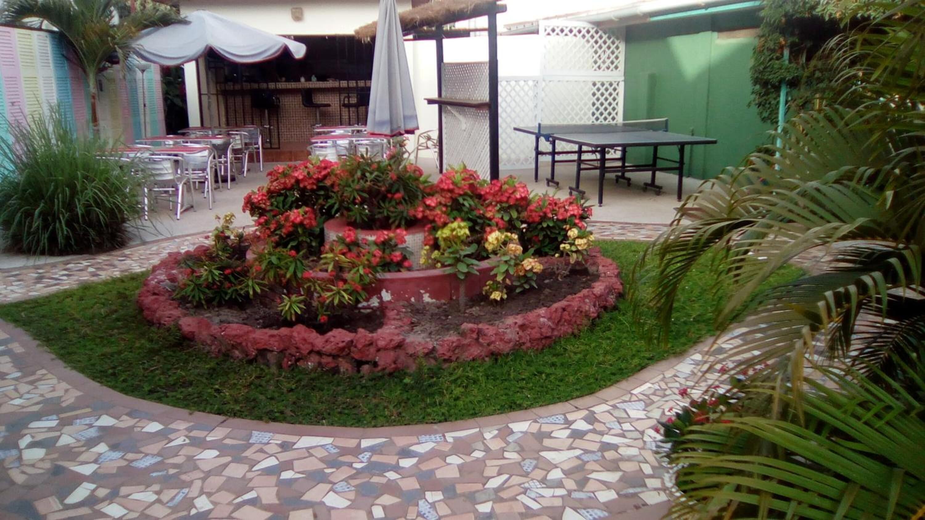 A patio area with a mosaic-tiled floor, a central flower bed with various plants, outdoor tables and chairs, and a pergola with a white canopy.