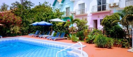 A swimming pool with lounge chairs and umbrellas, surrounded by lush greenery and a multi-story building with balconies.