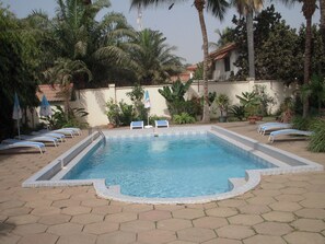 A rectangular swimming pool with a tiled edge, surrounded by lounge chairs and umbrellas, with palm trees and a building in the background. - Riyan Apartments (Serrekunda)