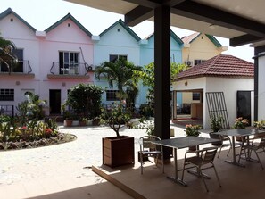 A covered outdoor patio with a table and chairs, surrounded by colorful buildings and potted plants. - Riyan Apartments (Serrekunda)