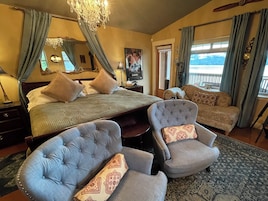 Un dormitorio con una cama, dos sillones, una araña, un cuadro y vistas al exterior.