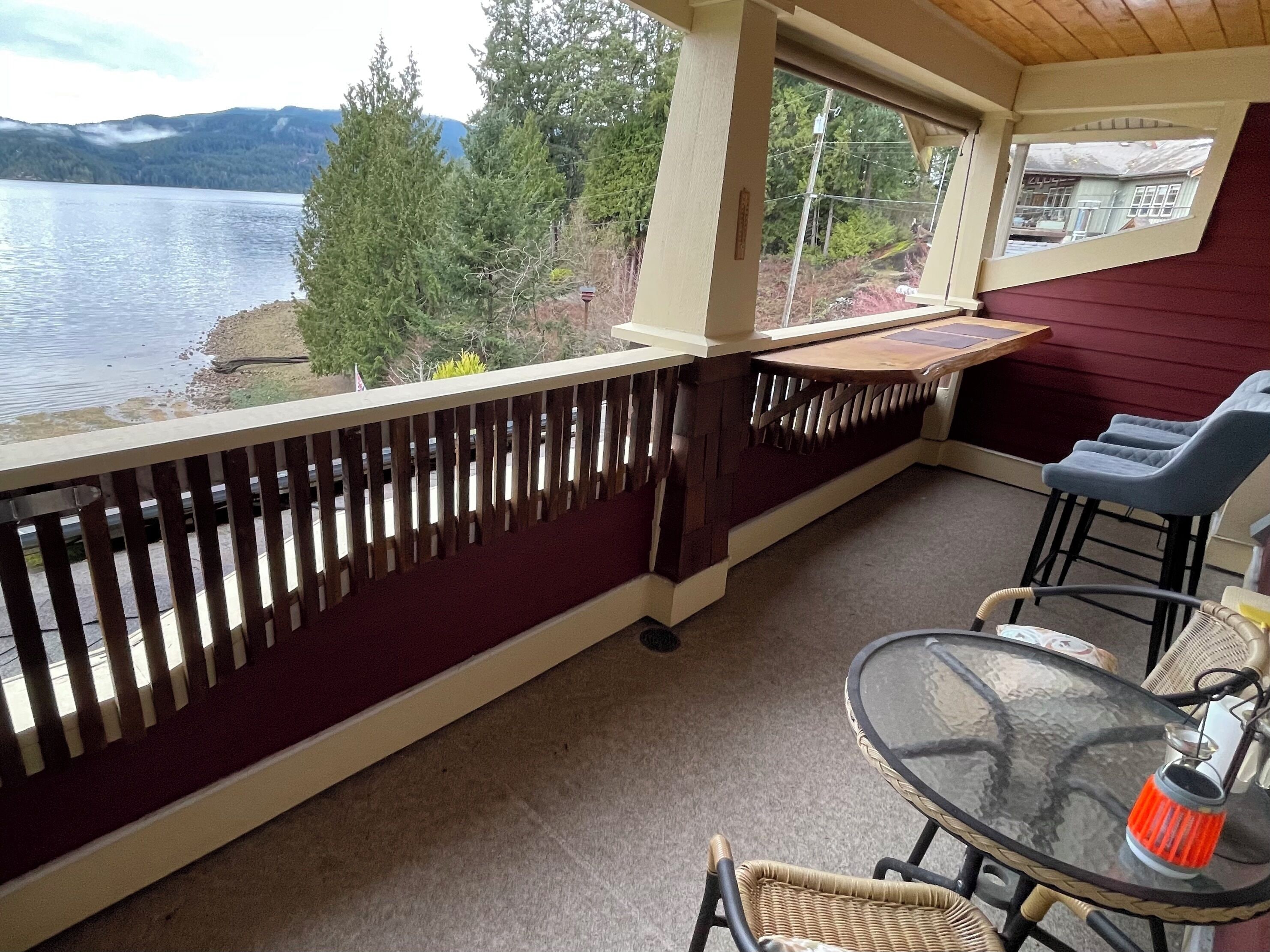 A covered outdoor seating area with a view of a lake and mountains, a glass table, wicker chairs, and a red wall.