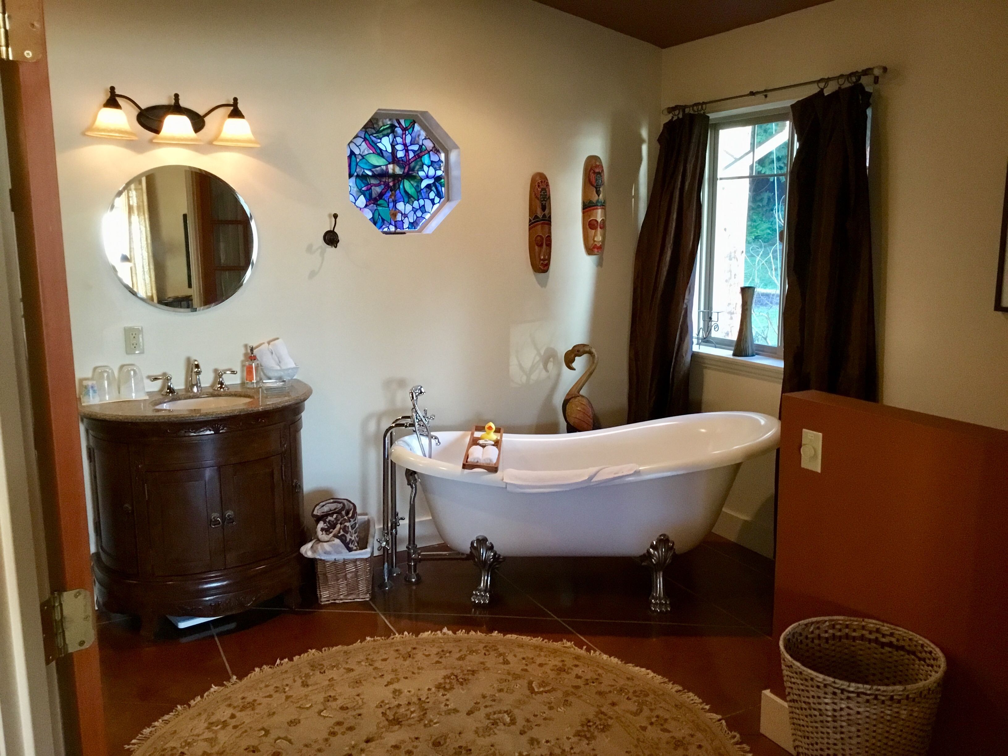 A bathroom with a freestanding bathtub, a round mirror, a stained glass window, and wooden wall decor.
