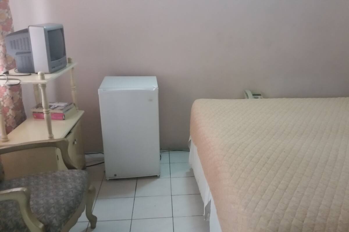 A bedroom with a bed, a desk with a computer, and a refrigerator.