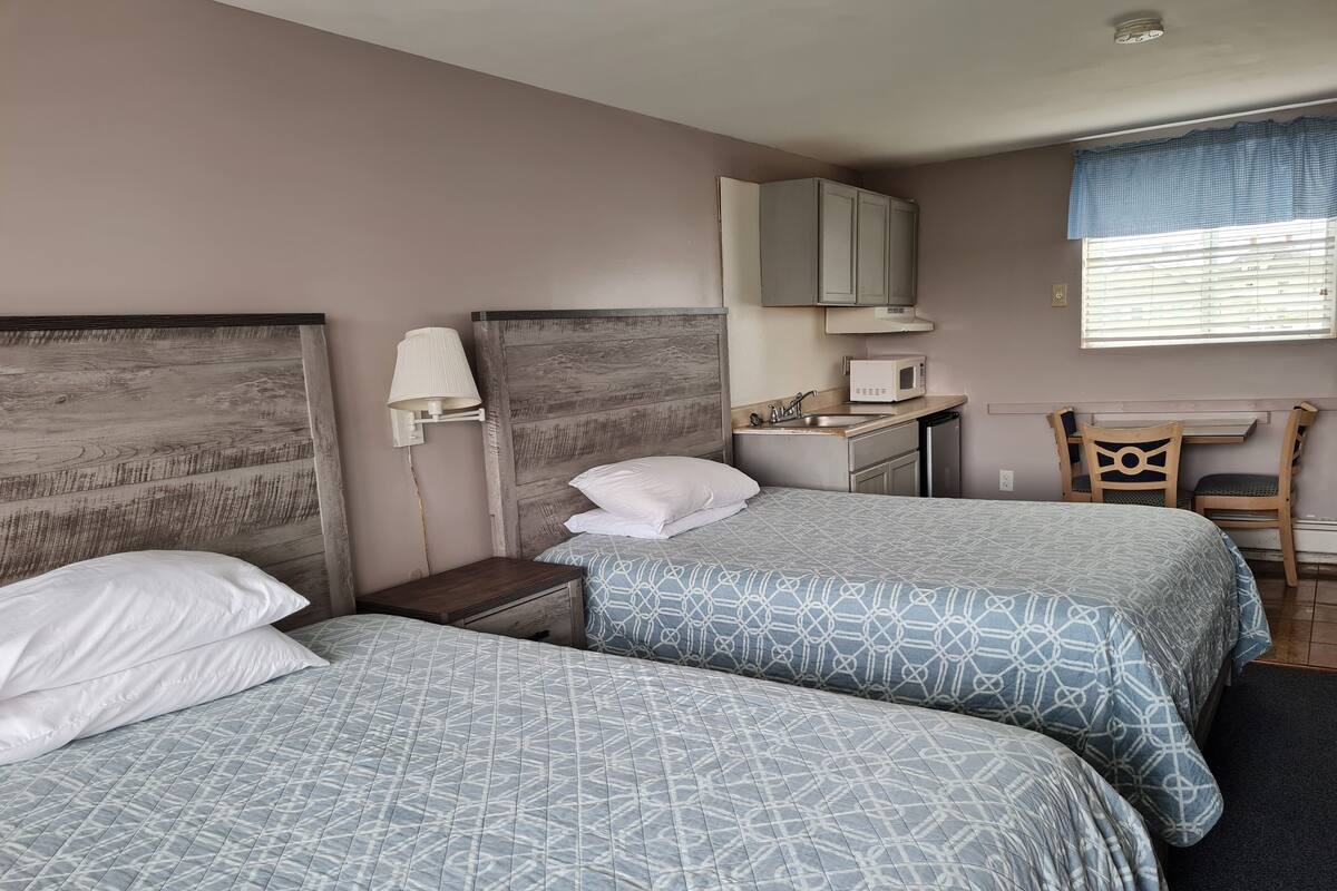 A bedroom with two beds, a nightstand, a desk with a chair, a microwave, and a window with blinds.