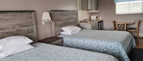 A bedroom with two beds, a nightstand, a desk with a chair, a microwave, and a window with blinds.