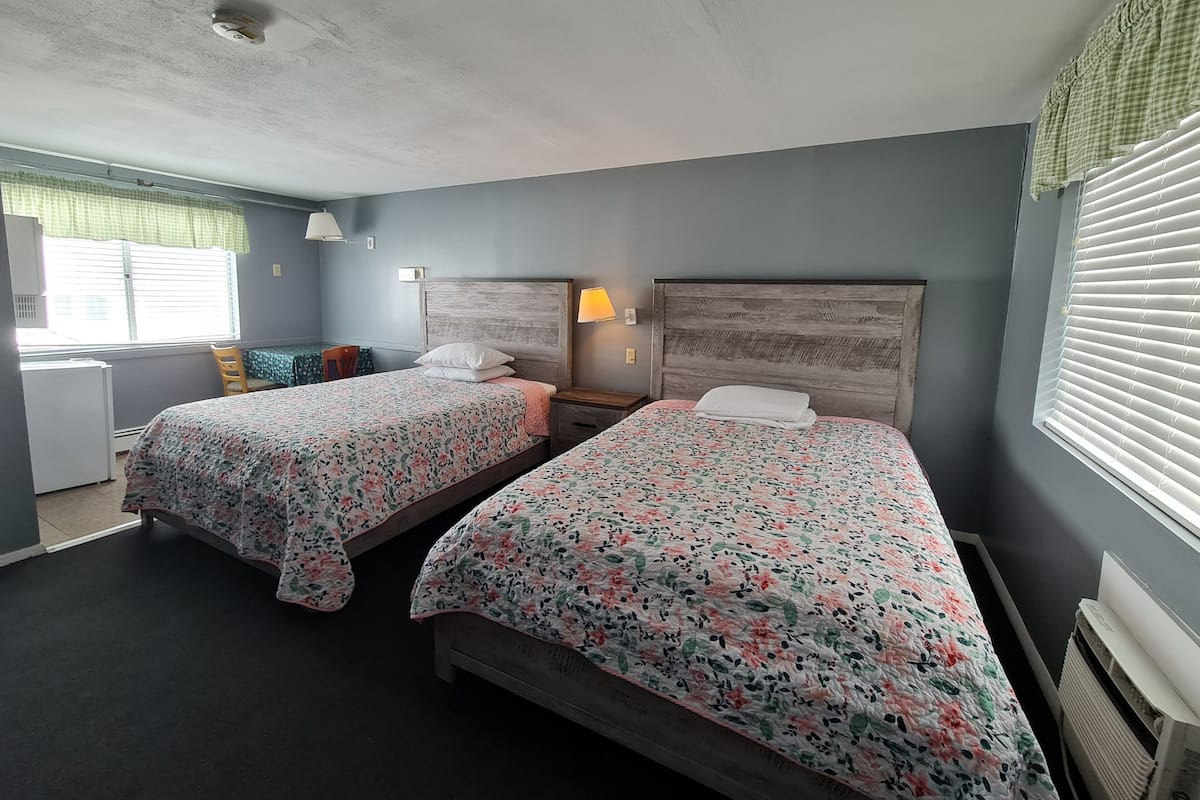 A room with two beds, each with floral bedding, a small table, a chair, a desk, and a window with blinds.