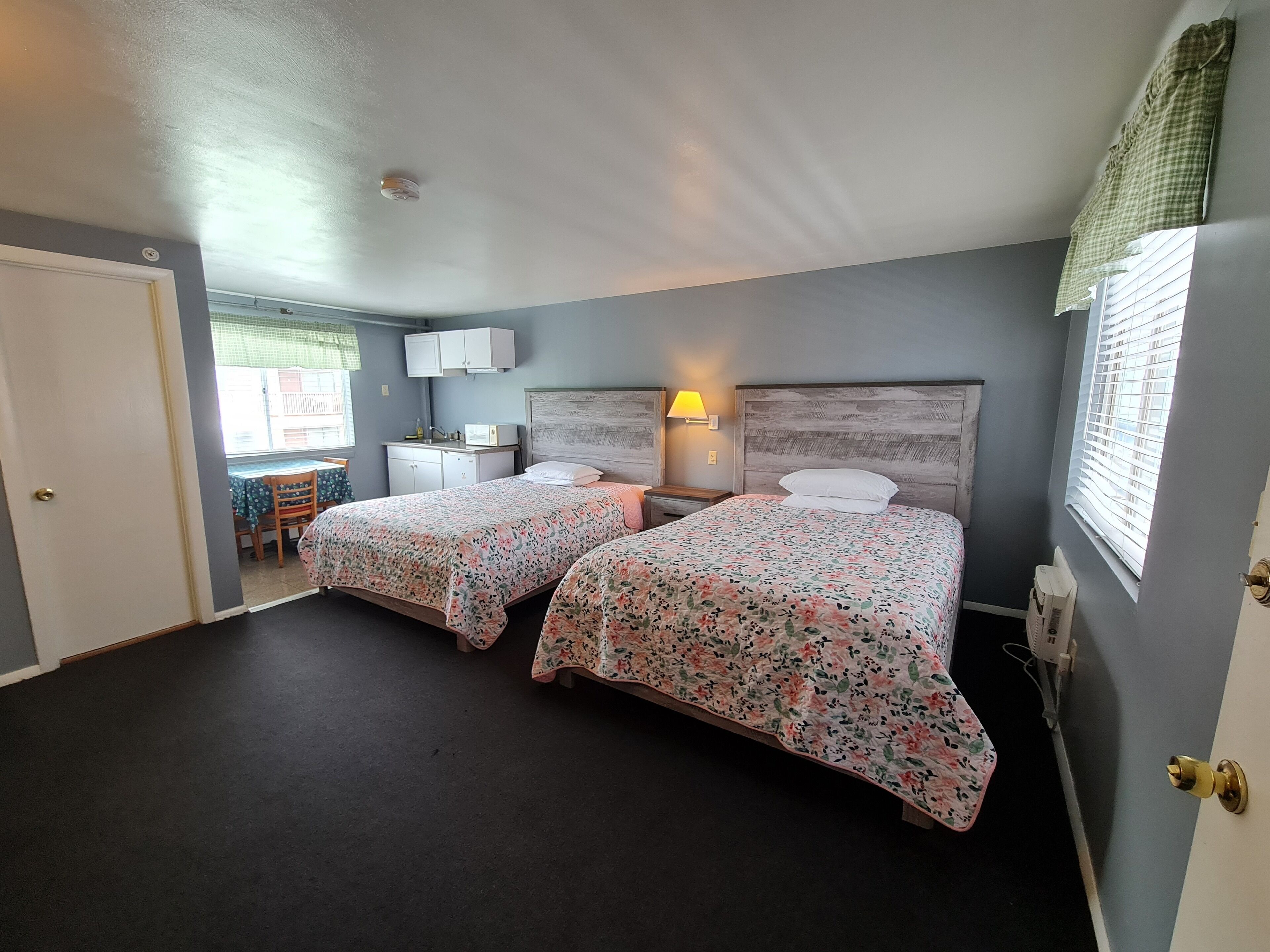 A room with two beds, a desk, a chair, a window with blinds, and a door.