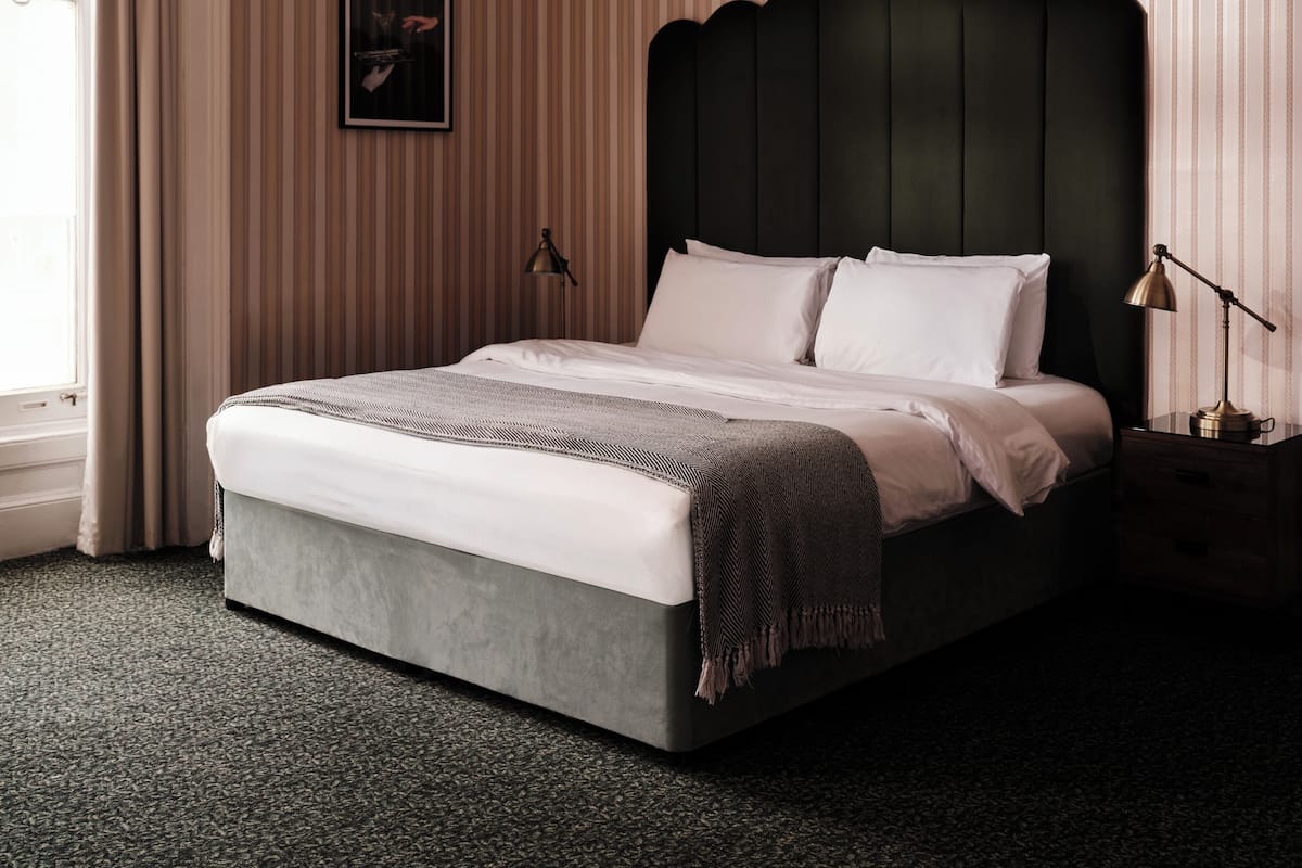A neatly made bed with a grey headboard, a bedside lamp, and a framed picture on the wall.