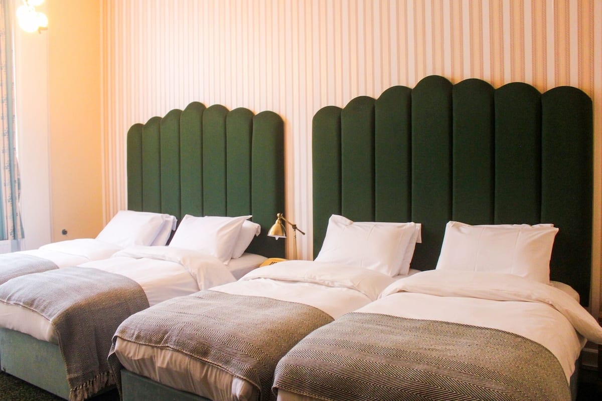 A hotel room with two single beds, each with a green headboard, white bedding, and a lamp on the nightstand.