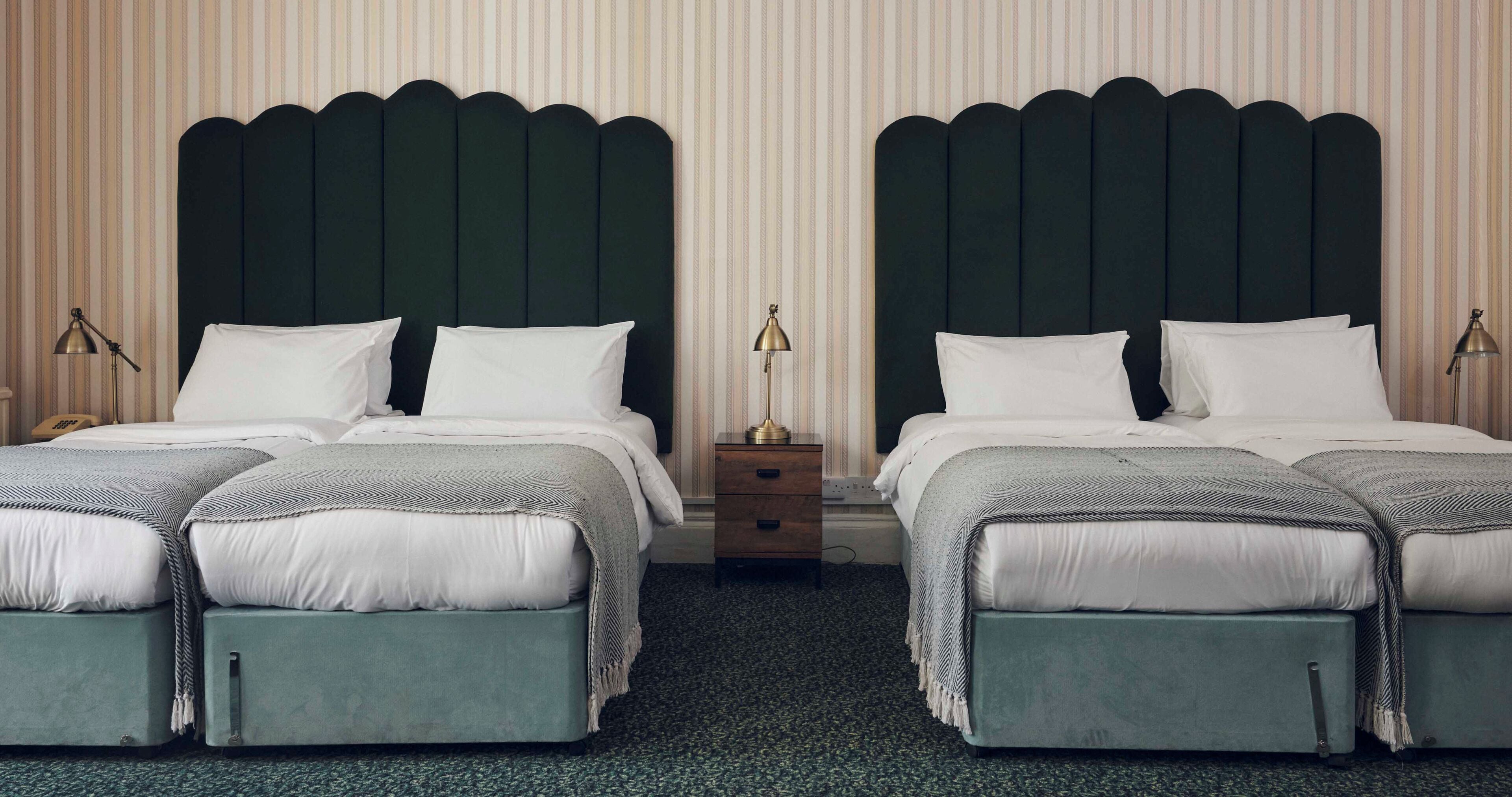 Ein Doppelzimmer mit zwei Betten, jeweils mit einem grünen Kopfteil und weißer Bettwäsche. An den Betten befinden sich Nachttische mit Lampen und eine kleine Schublade.