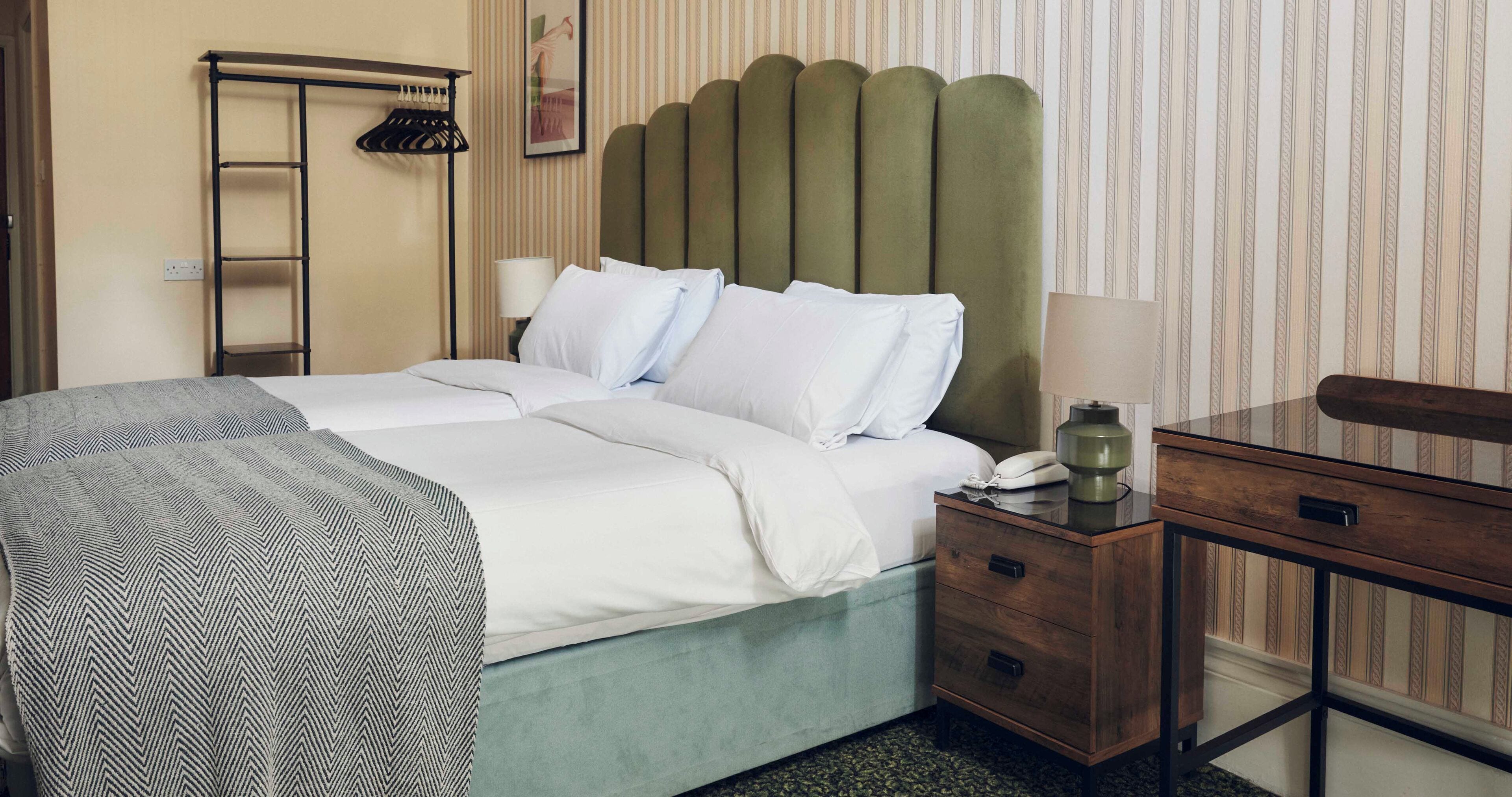 Un cuarto de hotel ordenado con cama, una mesita de noche con lámpara, un mesillón de madera y un guardarropa.