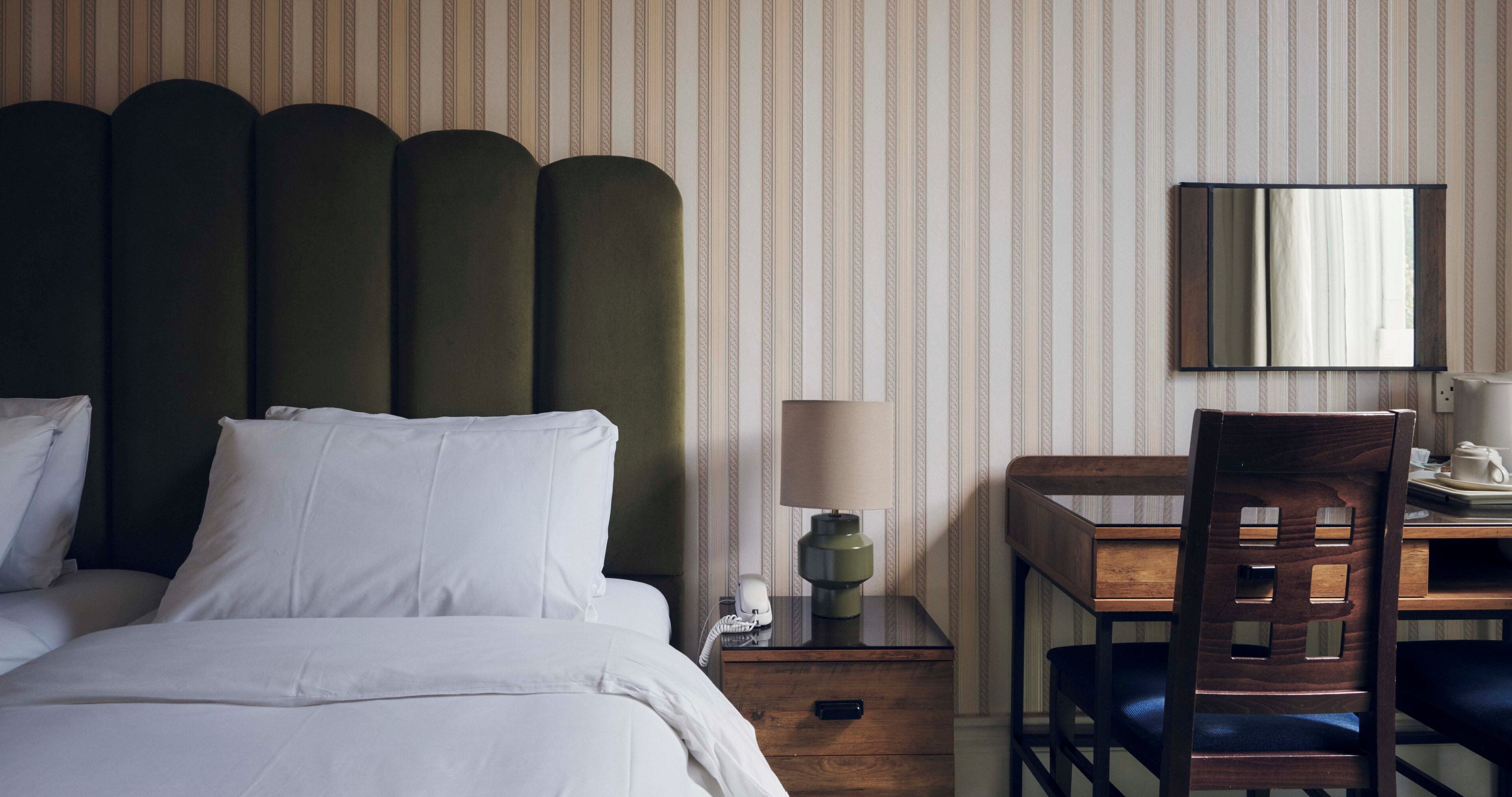 Ein Hotelzimmer mit Bett, Schreibtisch mit Stuhl, Nachttisch mit Lampe und einem Spiegel an der Wand.