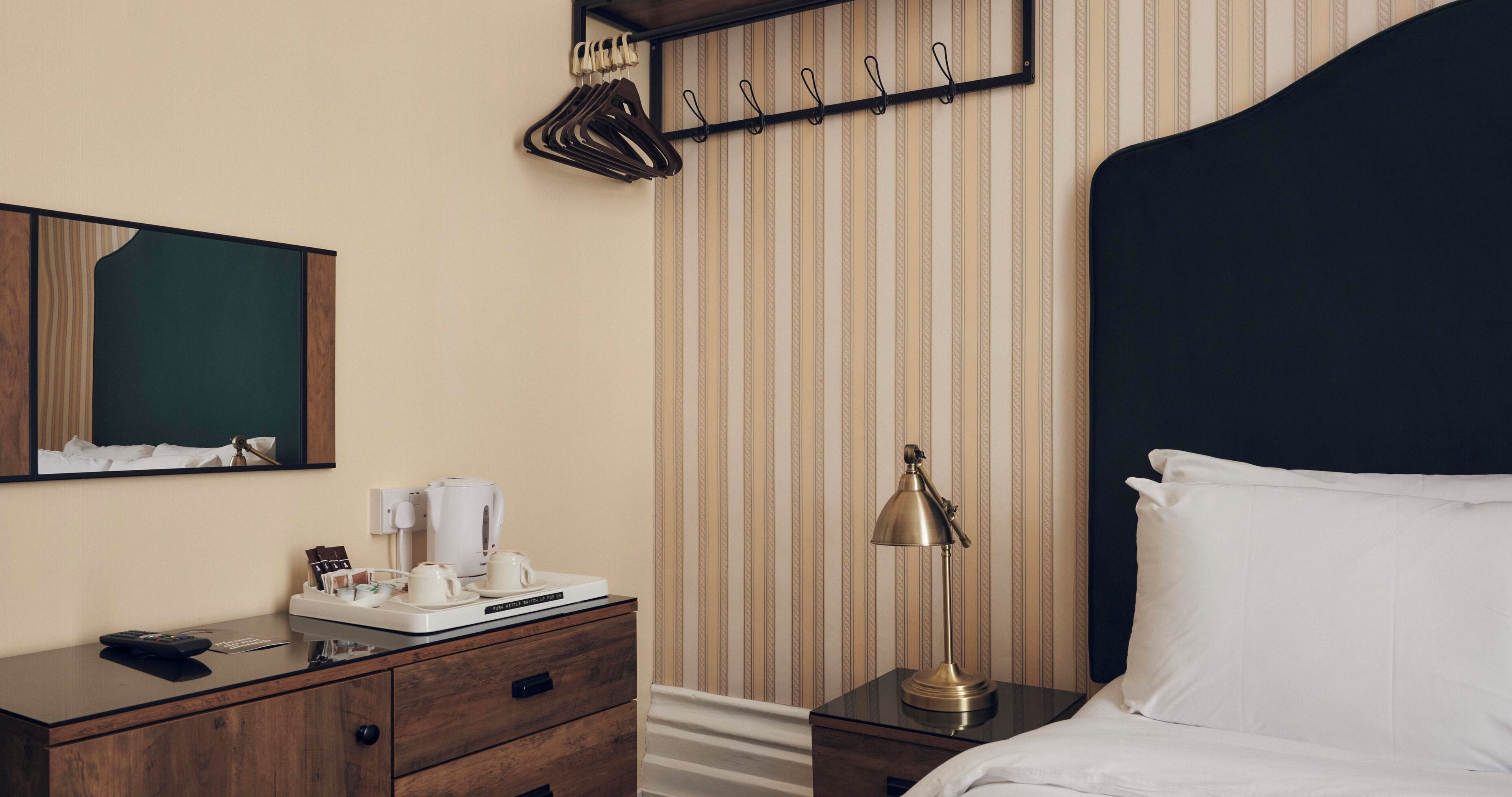 Habitación de hotel con tocador de madera, mesita de noche con lámpara, cama con sábanas blancas, perchero y un papel tapiz a rayas.