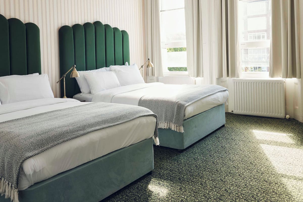A hotel room with two beds, a green headboard, white bedding, and a view of the city through large windows.