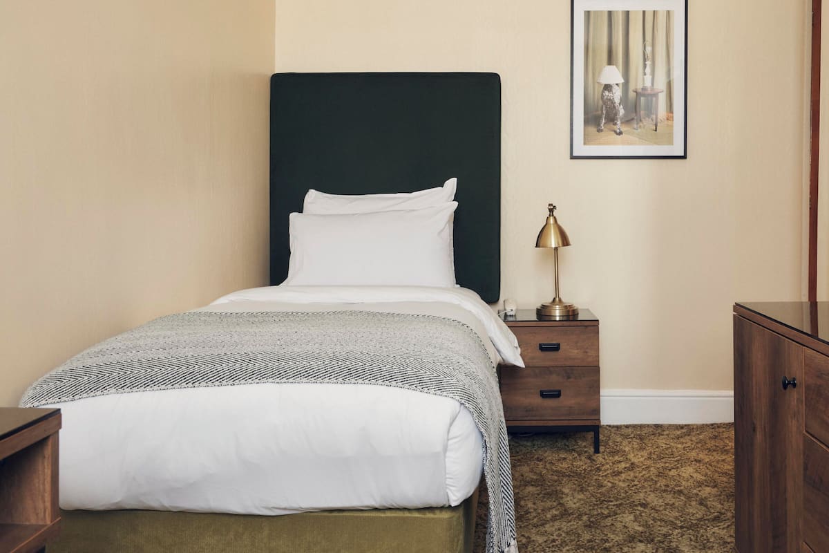 A neatly made bed with white linens, a dark headboard, a small bedside table with a lamp, and a wooden dresser with a mirror.
