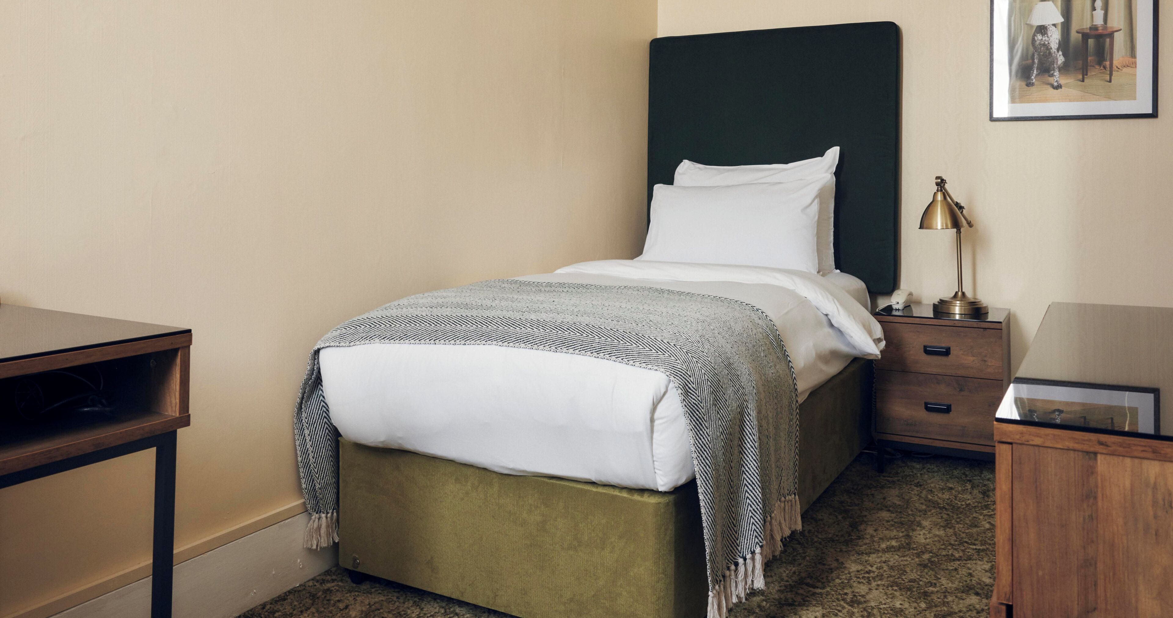 Un lit bien fait, avec une tête de lit rembourrée de couleur verte, une lampe de chevet et un tableau encadré accroché au mur.