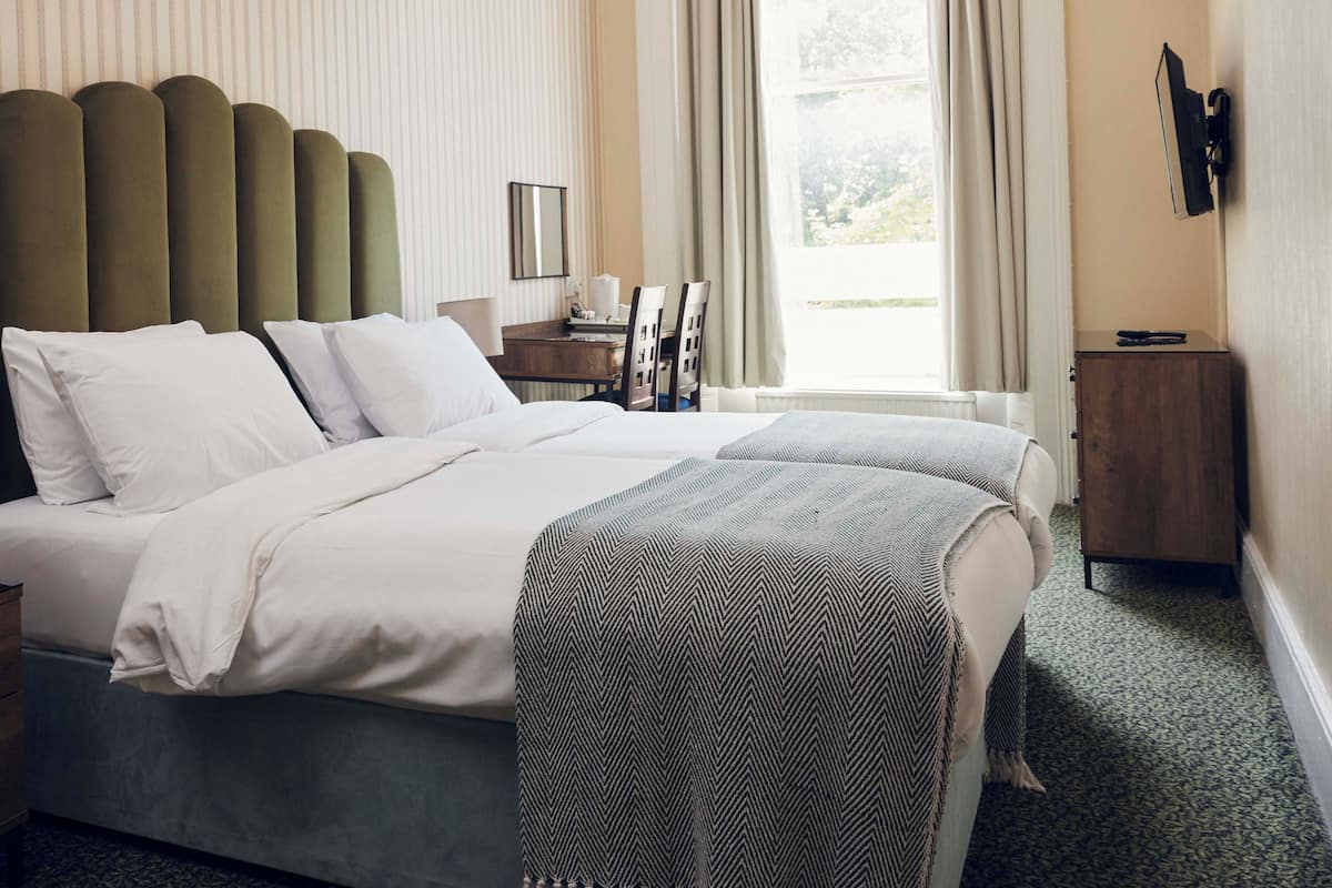 A hotel room with a large bed, a bedside table with a lamp, a desk with chairs, and a view of the outdoors through a window.