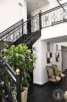 Una escalera con barandales negros ornamentados, una planta en maceta y dos sillones tapizados en un área de recepción.