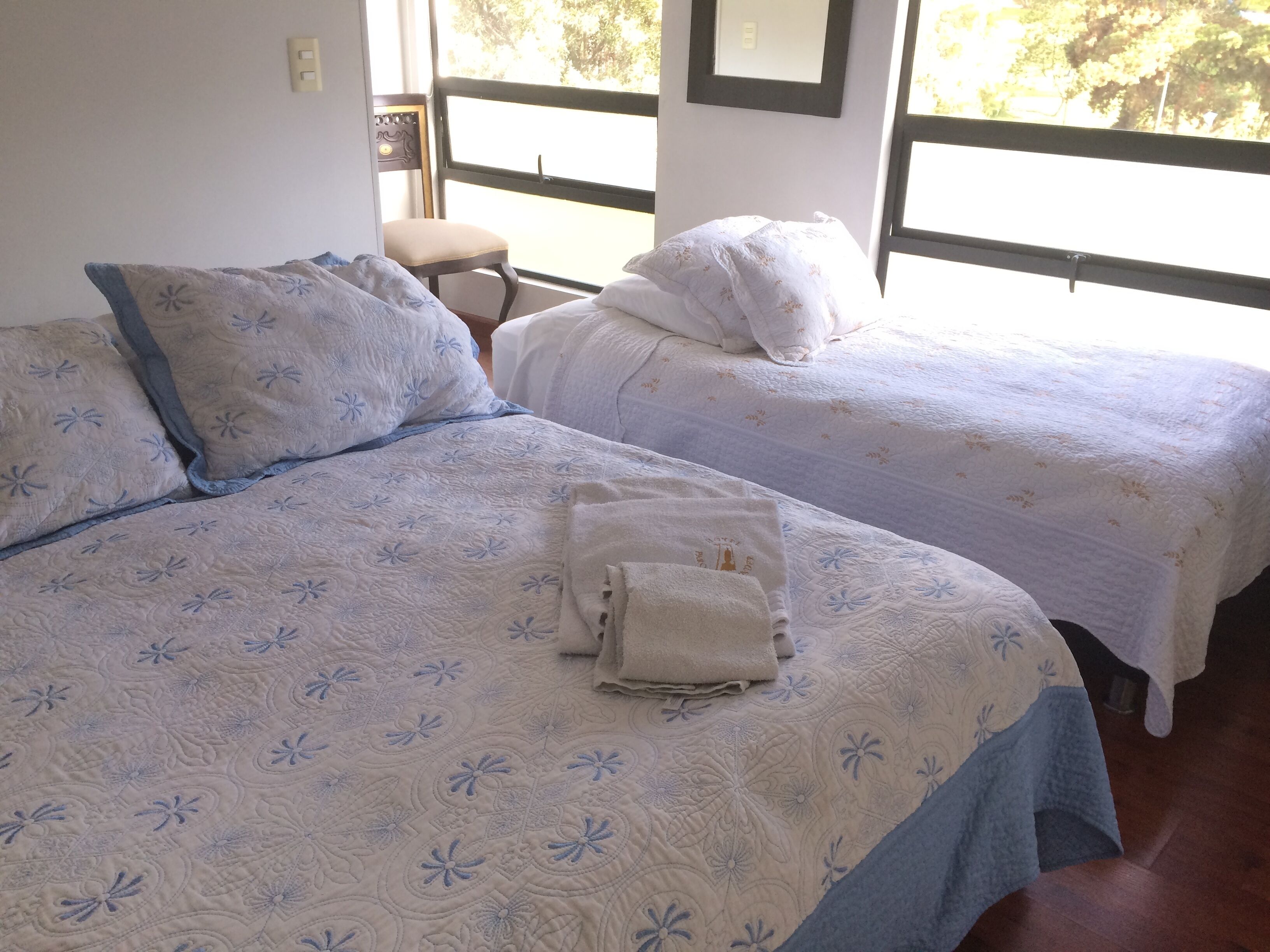 A hotel room with two beds, each with a floral-patterned bedspread and matching pillows. There is a chair and a small table in the corner.