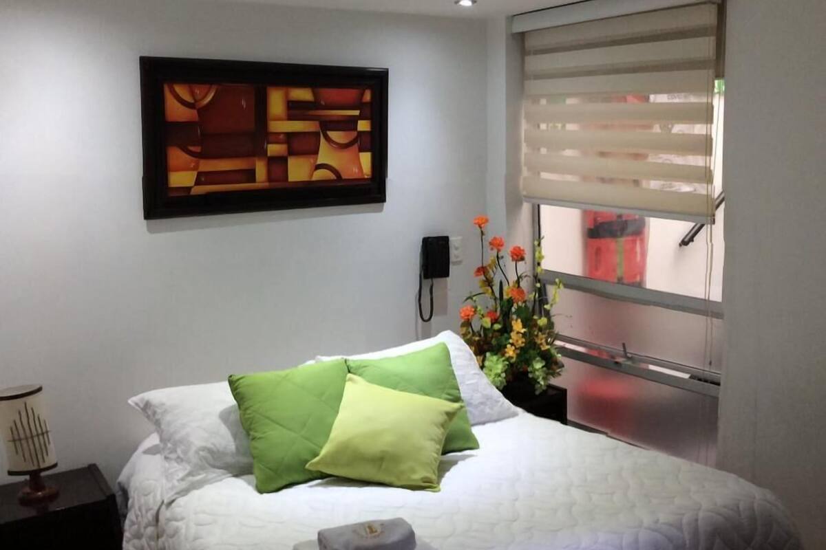 Una cama bien hecha con colcha blanca y almohadas verdes, una mesita de noche con lámpara, un cuadro abstracto enmarcado en la pared, y una ventana con persianas.