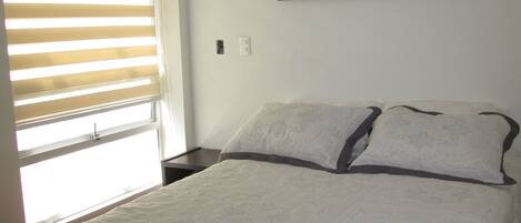 Un dormitorio con una cama, una ventana con persianas, una mesita de noche y un cuadro enmarcado en la pared.