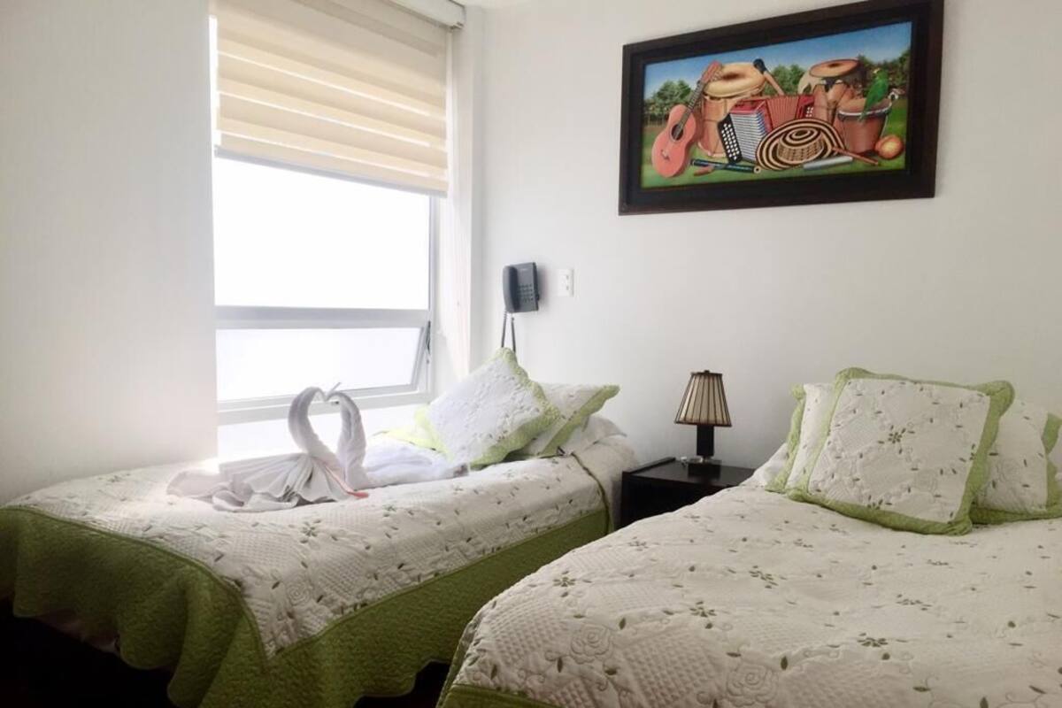 Habitación de hotel con dos camas, cada una con cubrecama verde y blanco, un cuadro sobre las camas y una ventana con persianas.
