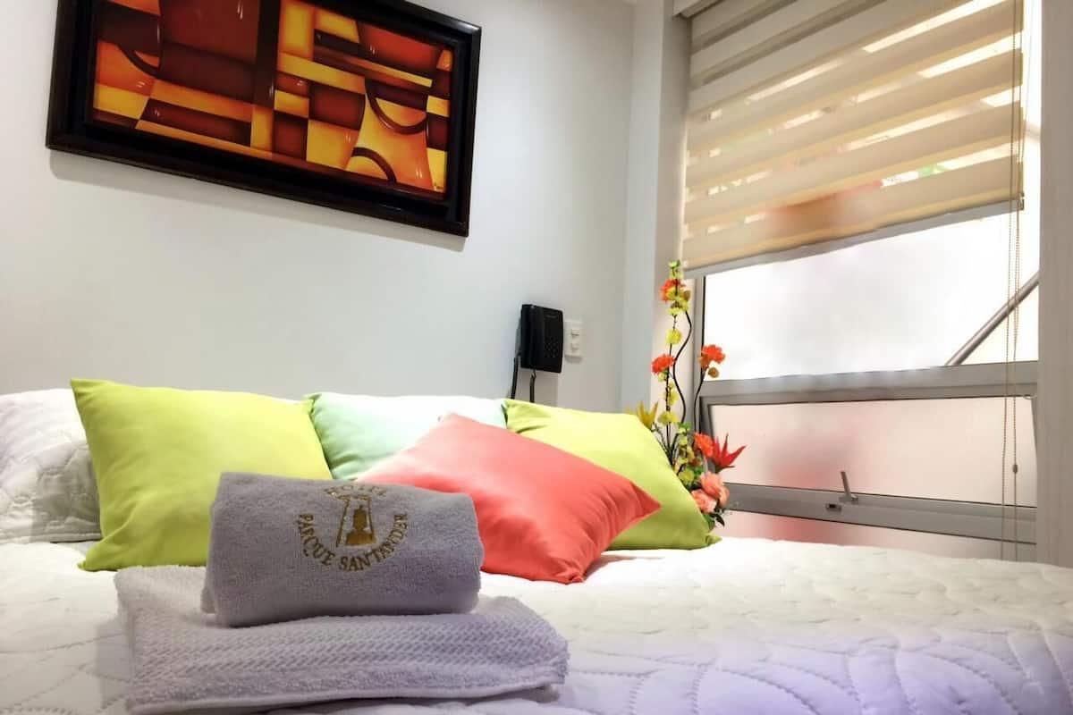 Una cama bien hecha con almohadas coloridas, un cuadro abstracto enmarcado en la pared y una ventana con persianas.
