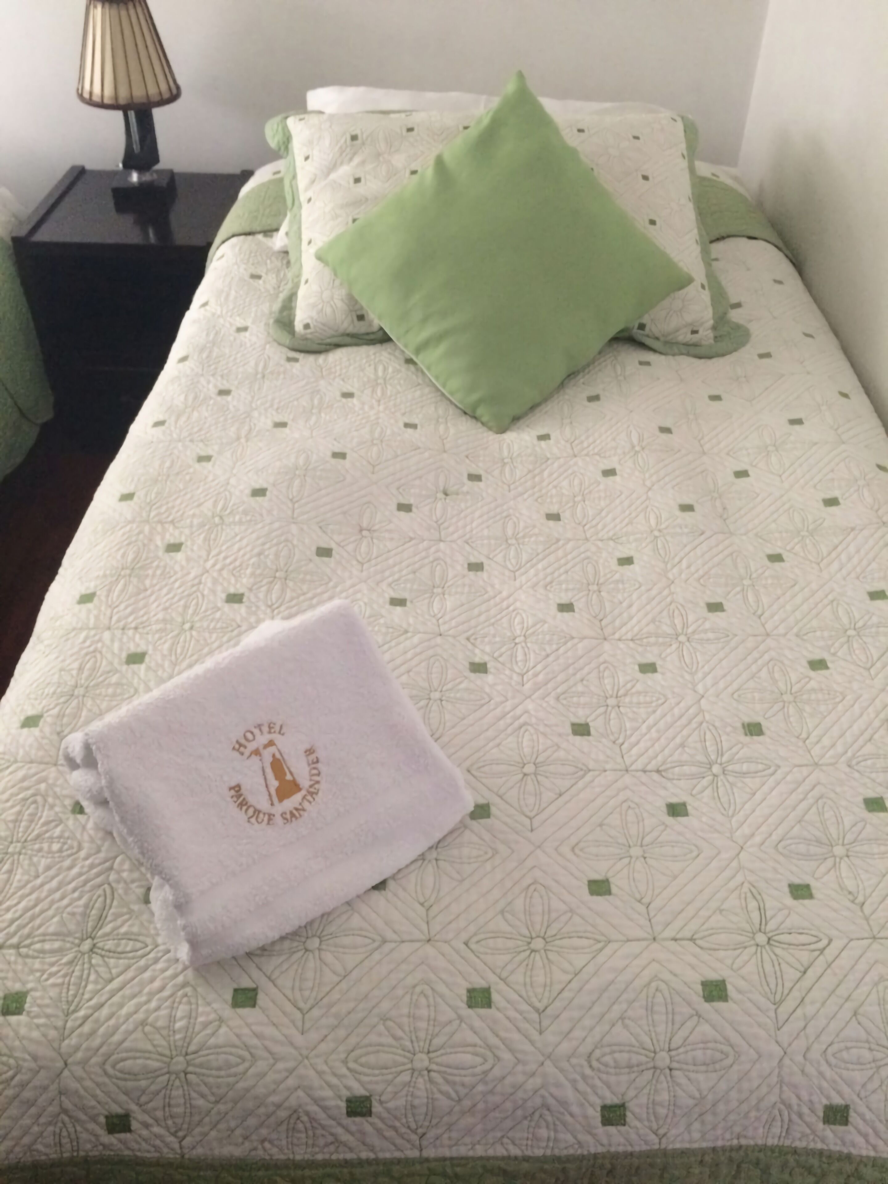 Ein ordentlich bezogenes Bett mit einem grünen Kopfkissen, einem weißen Handtuch mit Hotel-Logo und einer gemusterten Tagesdecke.