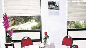 Un coin repas avec une table et des chaises, une fenêtre donnant sur un paysage verdoyant et un pont, ainsi qu’une affiche accrochée au mur.