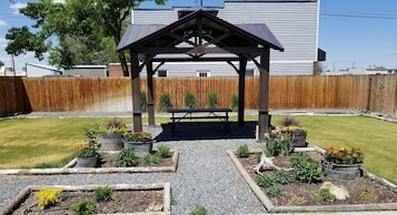 Uma pérgola de madeira com um banco em um jardim nos fundos, com canteiros de flores e cerca de madeira.