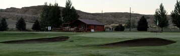 Um campo de golfe com bunkers, uma cabana de madeira e montanhas ao fundo.