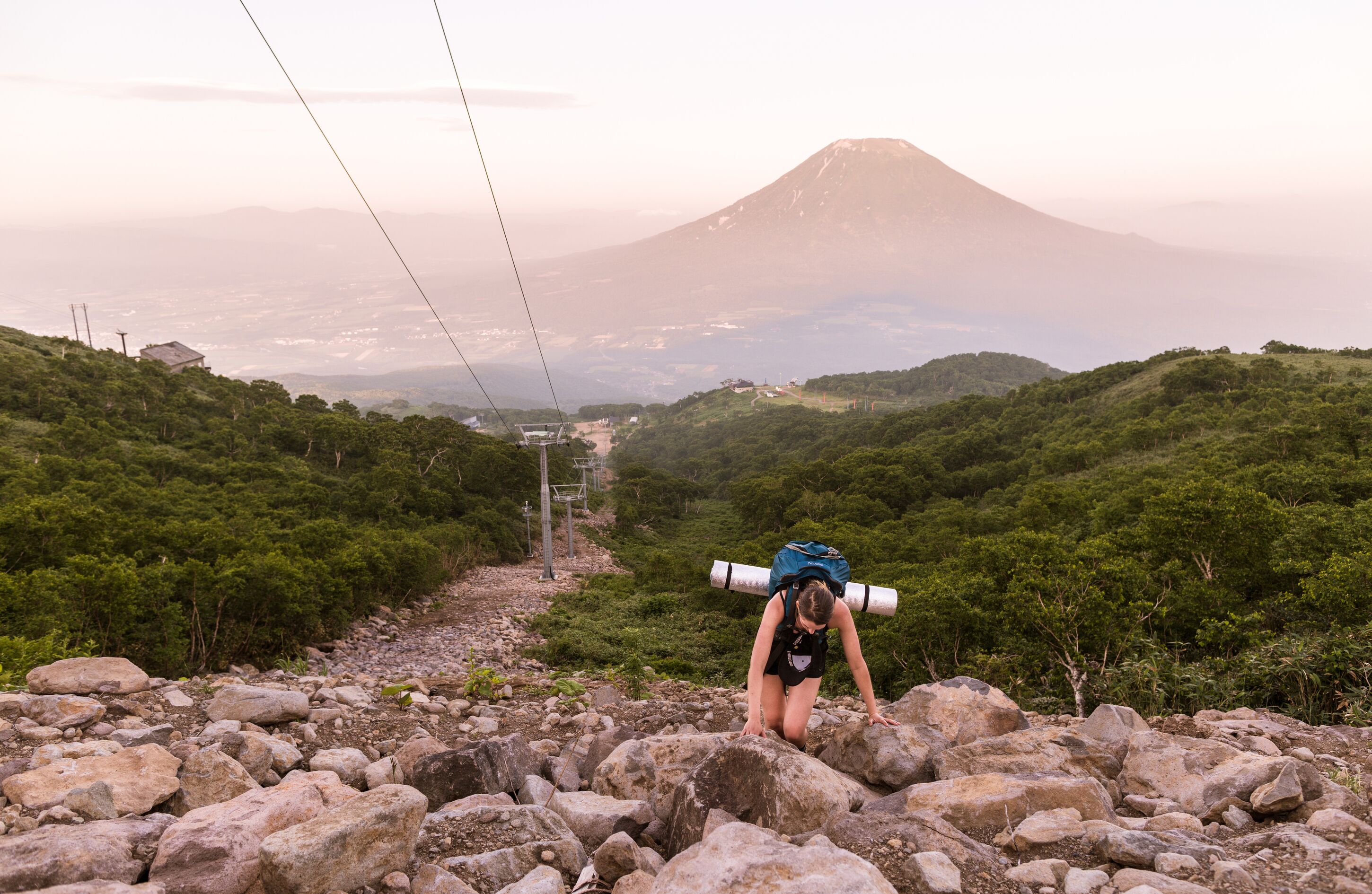 A hiker with a rolled-up mat on their back, navigating a rocky path with a mountain in the background.