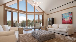 A spacious living room with a high ceiling, large windows, and a view of a mountain. There are two white sofas, a tufted ottoman, and a patterned rug. - The Orchards Niseko (Kutchan)