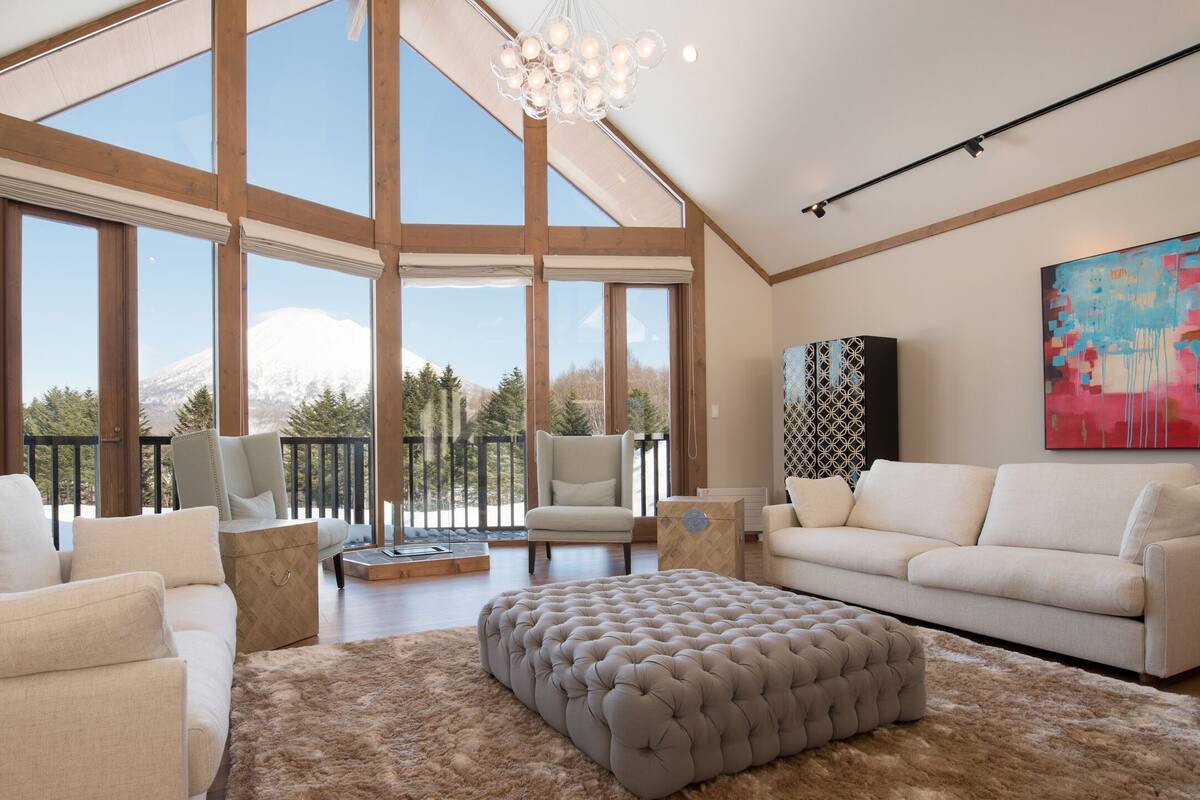A spacious living room with a high ceiling, large windows, and a view of a mountain. There are two white sofas, a tufted ottoman, and a patterned rug.