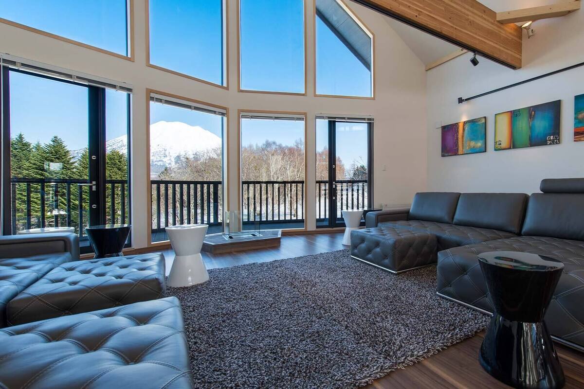 A spacious living room with a large sectional sofa, a coffee table, and a rug. There are large windows offering a view of trees and mountains.