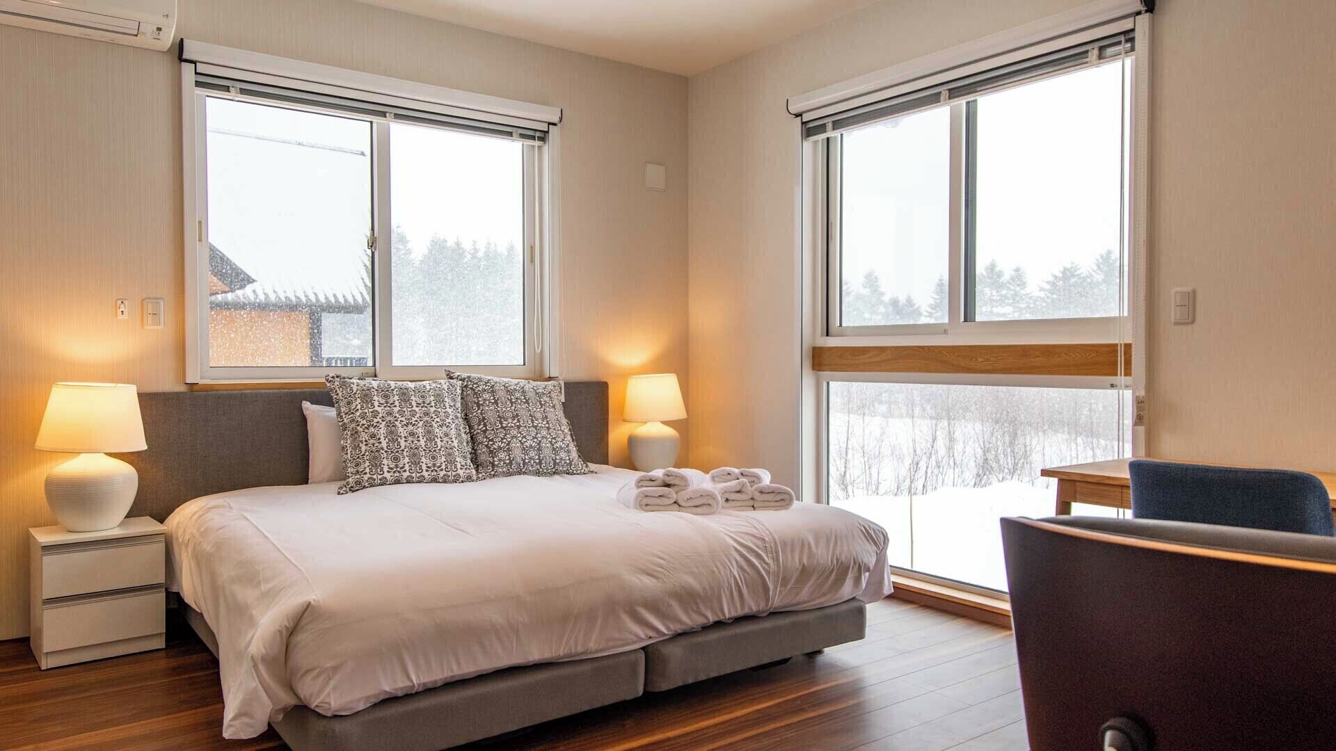 A hotel room with a bed, two bedside lamps, a chair, and large windows with a view of a snowy landscape.